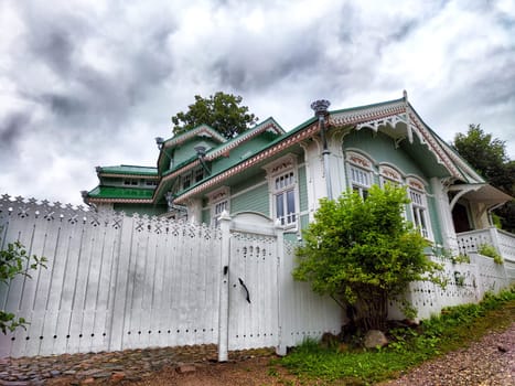 A charming wooden house featuring decorative elements stands behind a white fence, set against a backdrop of cloudy skies.