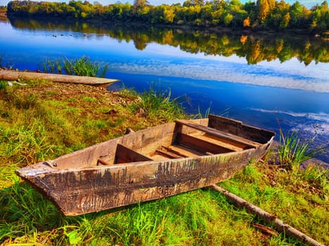 A weathered boat sits on the riverbank lush with green grass and vibrant fall colors reflecting on the water.