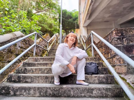 A woman relaxes on stairs surrounded by greenery, taking a moment to enjoy her travel experience. The adventures of middle-aged female tourist on a trip