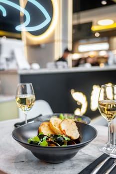 Mussels in cream sauce with croutons in a black plate on a table in a restaurant with a glass of white wine