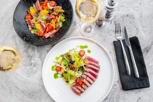 two plates with tuna tataki salad with lettuce, avocado and cherry tomatoes on a marble table in a restaurant next to glasses of white wine and cutlery