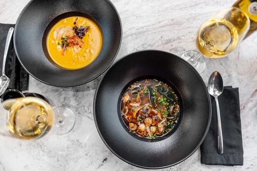 Two plates of soup: pumpkin cream soup with shrimp and miso soup with mushrooms on the table in a restaurant with a glass of wine