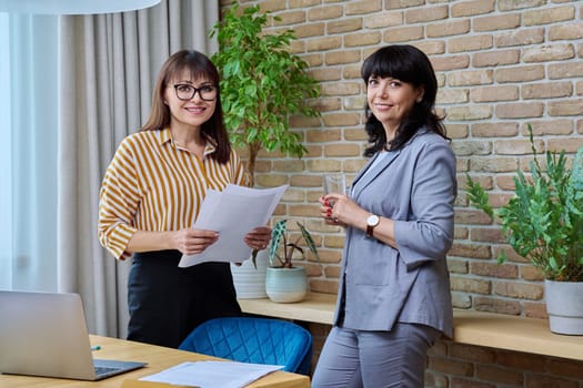 Two mature business women colleagues working together in office. Smiling posing females looking at camera holding business papers contracts. 40s 50s business people, work, job