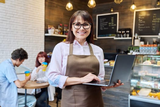 Portrait of middle aged woman owner of bakery coffee shop worker, smiling looking at camera female with laptop in her hands, inside cafeteria. Small business, entrepreneur, work, people concept