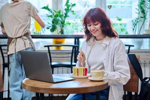 Female college student with laptop in cafeteria at table with cup of coffee and piece of cake. Internet online technology for leisure communication blogging learning chat, youth lifestyle concept