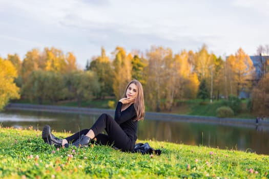 A beautiful girl poses while sitting on the grass by a pond in an autumn park