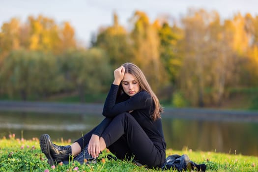 A beautiful girl poses while sitting on the grass by a pond in an autumn park