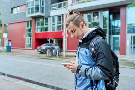 Fashionable young male with backpack using smartphone outdoors in city. Hipster with trendy hairstyle beard with phone in hands mobile applications for tourism travel communication entertainment study