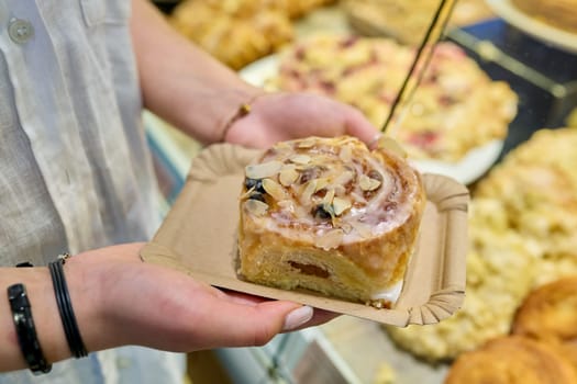 Bun with marzipan in hands, against the backdrop of display case in pastry bakery. Delicious fresh pastries, flour food, bakery cafeteria small business concept