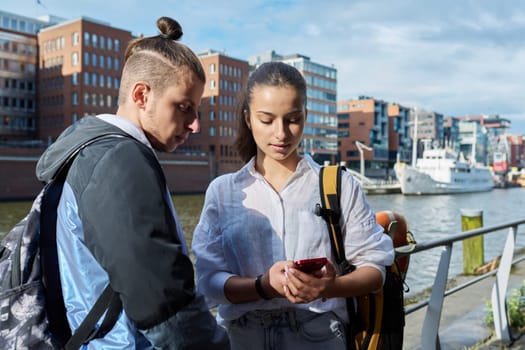 Teen friends guy and girl standing together holding smartphones looking at screen using mobile phones outdoor on city. Internet digital technology applications for leisure study communication