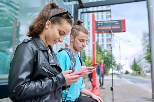 Teenage young people at city bus stop using smartphones, mobile applications for navigating transport timetables