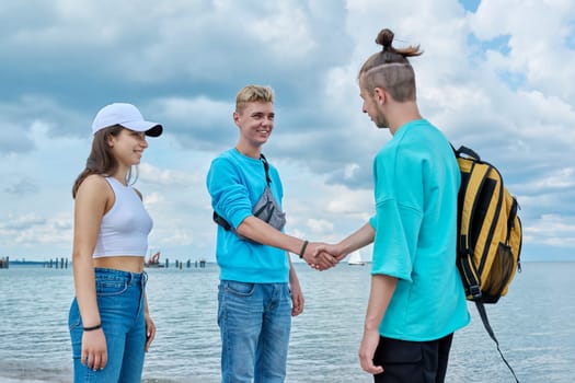 Young teenage friends guys and girl greet each other, shake hands, greeting, joy of meeting, outdoor on beach. Youth adolescence summer vacation communication leisure friendship fun lifestyle holiday