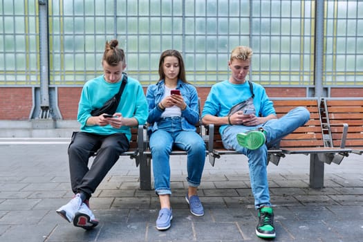 Group of teenage friends using smartphones sitting on chairs on train station platform. Youth, adolescence, digital technology, mobile applications, communication leisure learning social networks