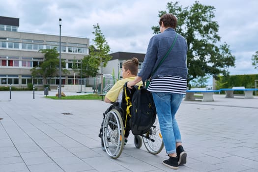 Back view of woman with boy in wheelchair near the school building, copy space. Children, disability, education, activity, health, lifestyle concept