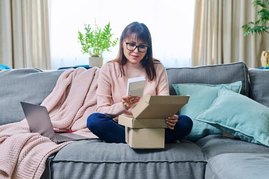 Mature woman sitting on sofa at home, living room, unpacking cardboard box with online shopping, buying new mobile phone smartphone. Internet shopping, postal delivery, delivery service