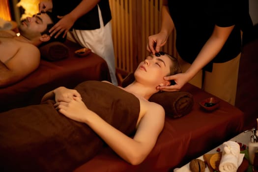 Couple customer enjoying relaxing anti-stress head massage and pampering facial beauty skin recreation leisure in warm candle lighting ambient salon spa in luxury resort or hotel. Quiescent
