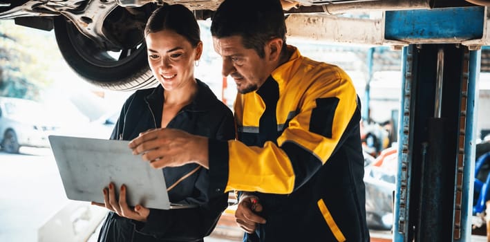 Two vehicle mechanic working together underneath lifted car, conduct car inspection with laptop. Automotive service technician in uniform carefully make diagnostic troubleshooting. Panorama Oxus