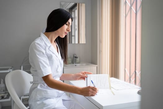 Female doctor filling up patient file. Young brunette therapist in white uniform sitting next to window. Female cosmetologist writing down patient information sitting at medical office