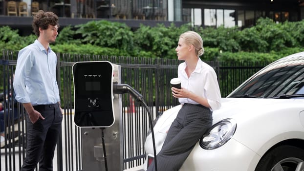 Progressive businessman and businesswoman leaning on electric car connected to charging station before driving around city center. Eco friendly rechargeable car powered by clean energy.