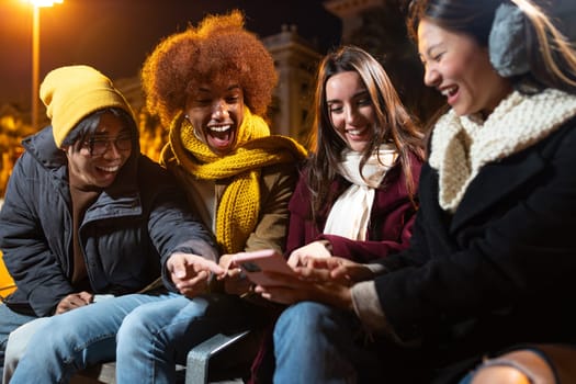 Happy multiracial friends looking at mobile phone together and laughing during winter night sitting on a city street bench. Technology and friendship concept.