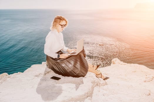 Successful business woman in yellow hat working on laptop by the sea. Pretty lady typing on computer at summer day outdoors. Freelance, travel and holidays concept.