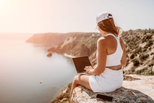 Digital nomad, Business woman working on laptop by the sea. Pretty lady typing on computer by the sea at sunset, makes a business transaction online from a distance. Freelance remote work on vacation