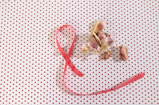 Pink ribbon next to garlic bulbs on a red polka dot backdrop, symbolizing culinary simplicity with visual charm.