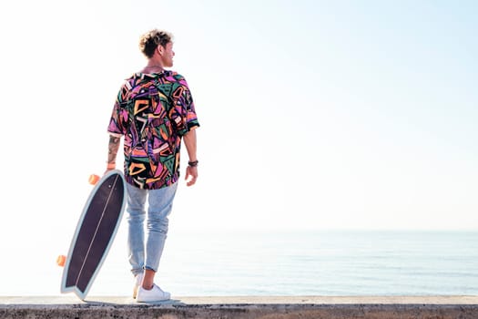 back view of young hipster male skater in trendy summer outfit carrying skateboard while standing on embankment against sea and cloudless sky in summer day