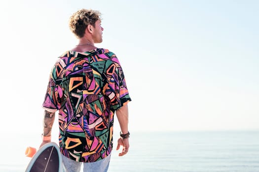 back view of young hipster male skater in trendy summer outfit carrying skateboard while standing on embankment against sea and cloudless sky in summer day