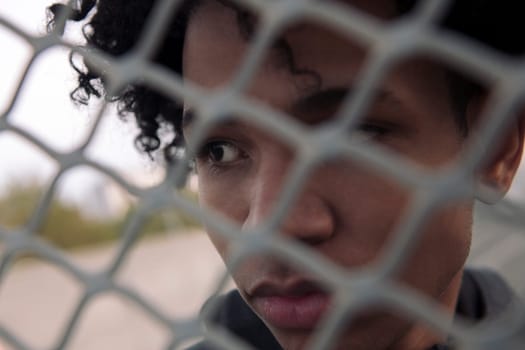 close up portrait of thoughtful teen african american male with curly hair looking away while standing behind metal chain link fence on urban street, concept of adolescence and mental health