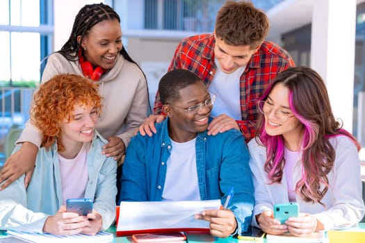 A group of five diverse students studying and collaborating at a library table working on a shared project, with books and devices on the table.