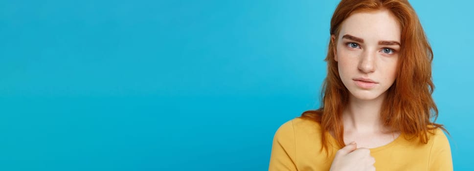 Headshot Portrait of tender redhead teenage girl with serious expression looking at camera. Caucasian woman model with ginger hair posing indoors.Pastel blue background. Copy Space