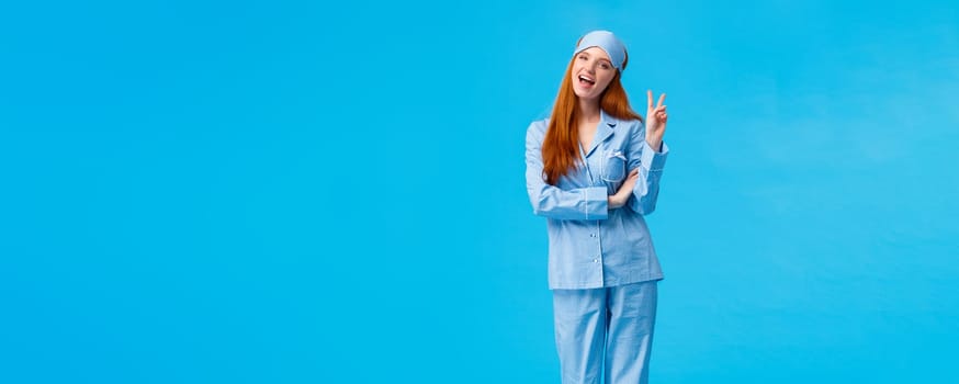 Saying cheeze. Cheerful and cute redhead upbeat girl in glamour pyjama, fancy nightwear having sleepover showing peace sign and smiling excited, having upbeat lucky morning, standing blue background.