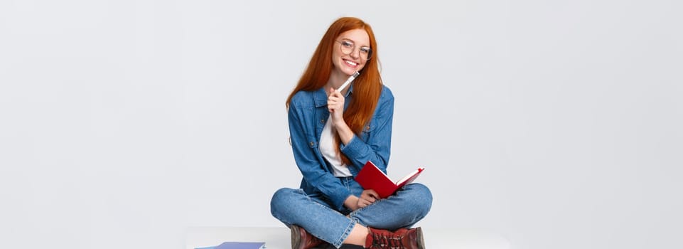 Charismatic, friendly cute redhead girl dreaming, taking notes or prepare homework while sitting on floor with legs crossed, holding notebook and pen, writing over white background.