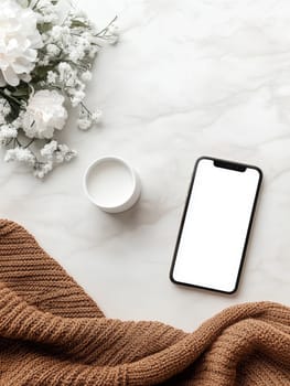 Elegant flat lay of a smartphone, white cup, and delicate flowers on a marble surface with cozy knit fabric, perfect for social media.