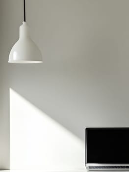 Clean, minimalist workspace featuring a laptop and white pendant lamp, with dramatic shadow play on a blank wall, perfect for copy space.