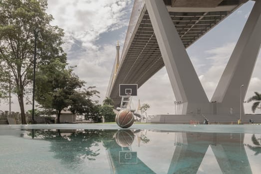 Bangkok, Thailand - 28 Sep, 2024 - Basketball ball on the Old cement court floor with the reflection of them on puddle of rainwater at Outdoor park with Suspension bridge and Sky in the background. Space for text, Selective focus.