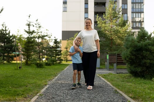 A joyful mother and her son stroll hand in hand along a gravel path in a park, surrounded by trees and laughter, sharing a moment of love and connection.