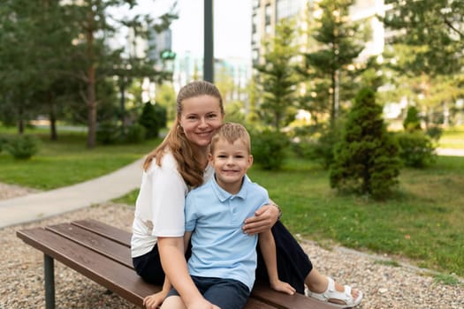 A mother lovingly embraces her young son as they sit together on a bench in a vibrant park. The setting sun highlights their warm relationship and laughter.