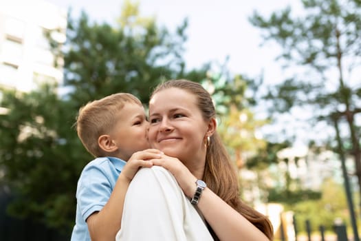 A mother smiles warmly as her son cuddles close, sharing a tender moment outdoors. Their bond shines brightly under the sun in a vibrant park setting.