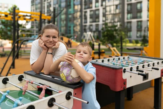 A mother and her son bond at a vibrant playground, with the boy holding a soccer ball and both of them smiling while enjoying a friendly game of foosball.
