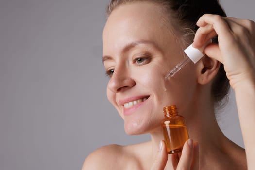 Happy smiling woman applying serum on her face, close eyes with pleasure. Redhead girl holding dropper with skin care product for healthy and glowing skin tone, blue background