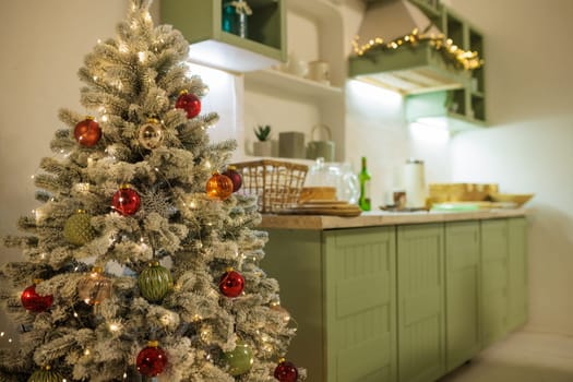 A cozy, bright kitchen decorated for Christmas with green cabinets, a small Christmas tree with decorations, and a festive table with snacks.