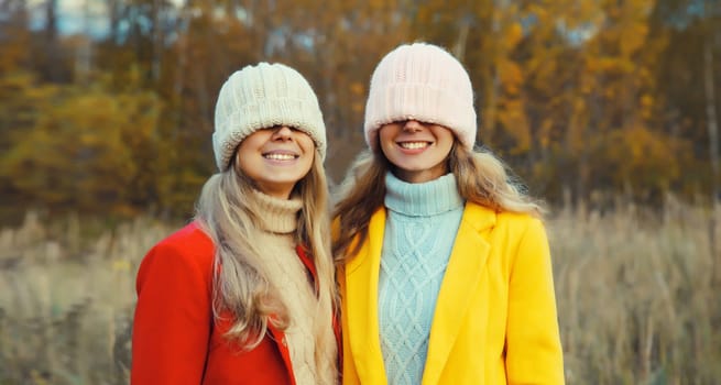 Happy two women friends pulling knitted winter hat over her face covering her eyes, funny girlfriends having fun together outdoors in park