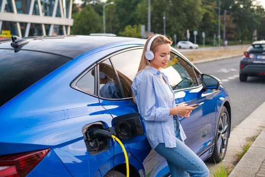 Woman listening to headphones while charging her electric car in an urban area during the day