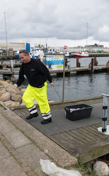 hanstholm,denmark,18-08-2024:A man in bright safety attire Fisherman taking the fish from the boat. Overcast skies and boats in the background create a typical maritime scene.