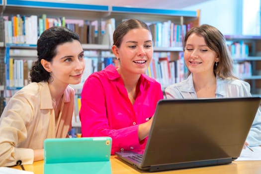Three young women sit in a library, working together with a laptop and tablet They are surrounded by bookshelves, suggesting a collaborative learning or study session in a relaxed academic environment