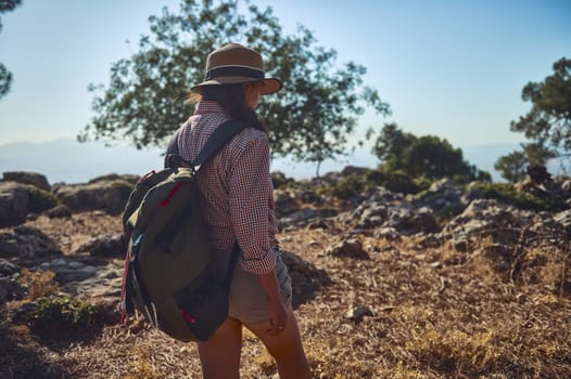 A woman with a backpack and hat hikes through a picturesque mountain landscape under a clear blue sky. Surrounded by lush greenery and rocks, she embraces the tranquility of nature.