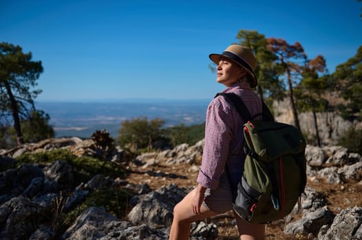 A woman enjoys a hike through rocky terrain, carrying a backpack and wearing a sun hat. The clear blue sky and expansive view highlight the adventurous spirit of outdoor exploration.