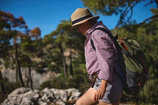 A woman wearing a hat and carrying a backpack hikes through a sunlit forest. The outdoor setting and casual attire suggest a relaxing and adventurous experience in nature.
